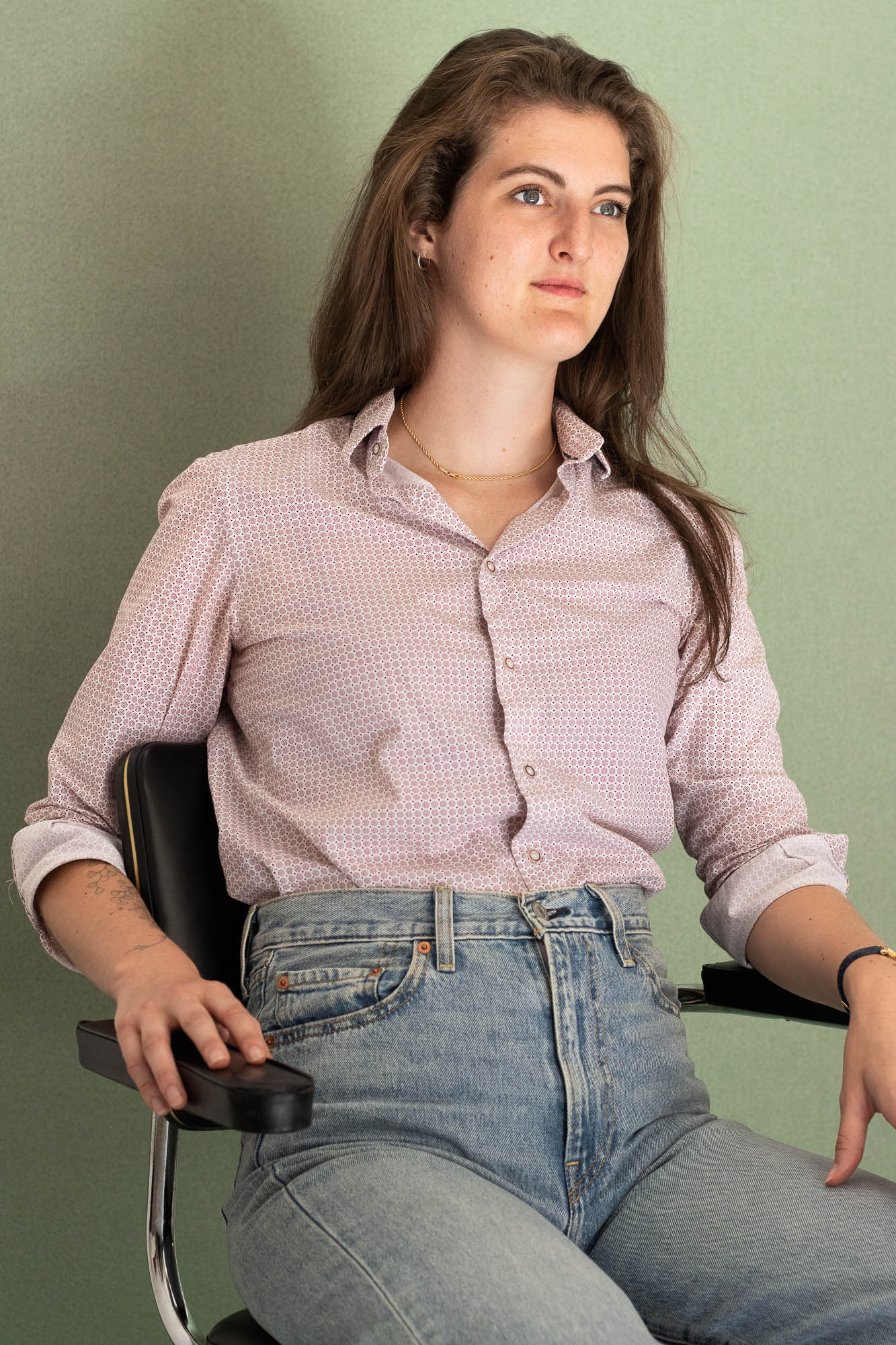 Portrait de Célestine Merckx assise sur une chaise inox avec un regard décidé, sur fond vert uni